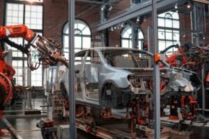 Orange KUKA robotic arms welding a bare metal car body on an automotive assembly line in a brick-walled factory hall