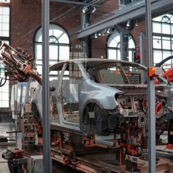 Orange KUKA robotic arms welding a bare metal car body on an automotive assembly line in a brick-walled factory hall
